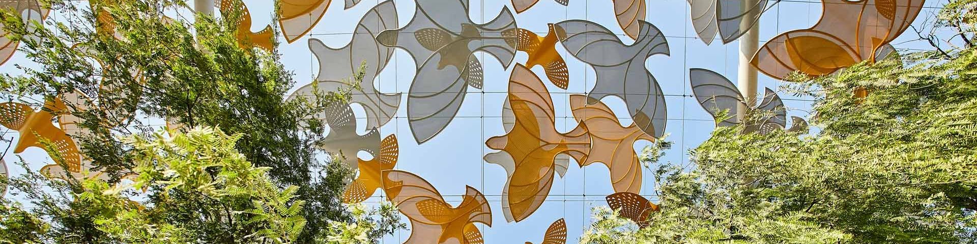 Cut out orange and grey shapes of birds, against a blue sky, with trees on either side. This is the view from beneath of a section of the Terra Sustainability Pavilion at Expo Dubai 2020.